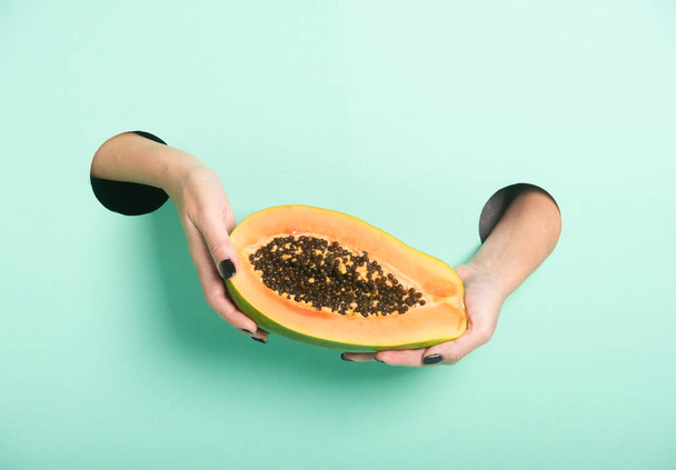 Female hands hold fresh papaya through a hole on neon mint background. Minimalistic creative isolated concept. - Foto, Imagen