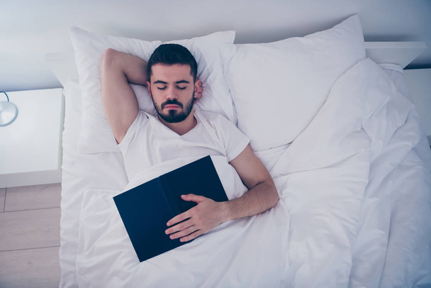 Top above high angle view portrait of his he nice attractive bearded guy lying on white bed resting sleeping peacefully fallen asleep holding in hand book at night late evening home room flat indoors - Foto, afbeelding