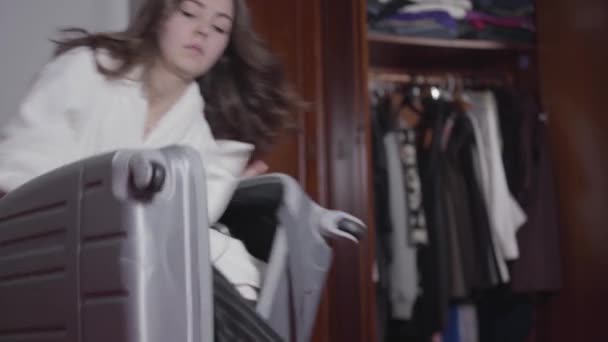 Beautiful hurrying Caucasian girl packing stuff in travel bag and leaving. Young brunette woman in white bathrobe late for flight. Tourist, traveler, lifestyle. - Imágenes, Vídeo