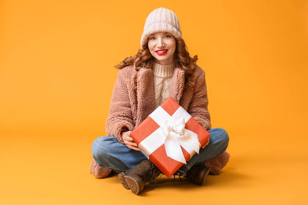 Happy young woman in winter clothes and with Christmas gift on color background - Foto, Imagem