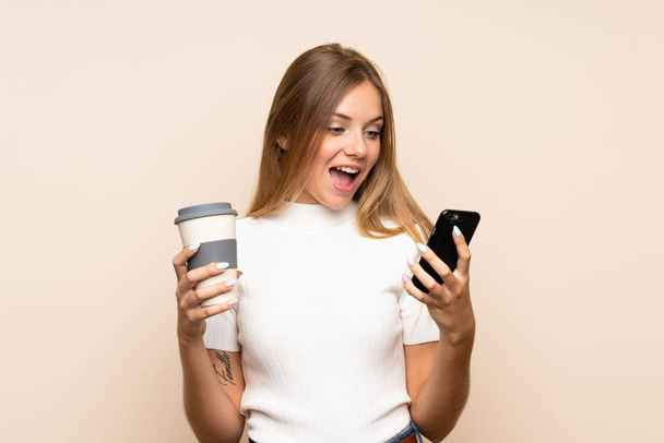 Młoda blondynka na odosobnionym tle trzymając kawę do zabrania i telefon - Zdjęcie, obraz