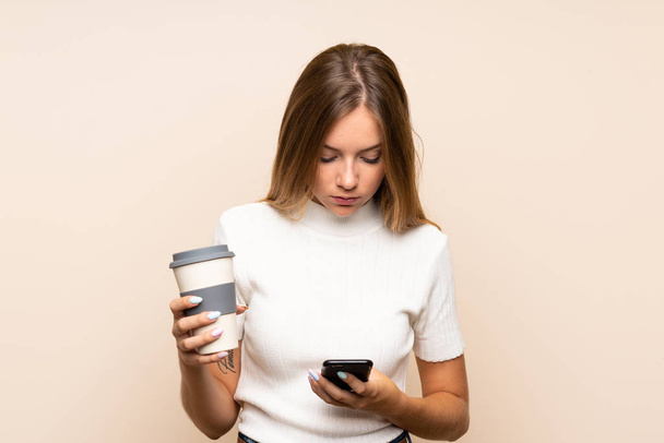 Giovane donna bionda su sfondo isolato in possesso di caffè da portare via e un cellulare
 - Foto, immagini