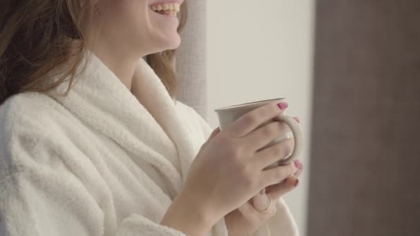Close-up of unrecognizable young Caucasian woman in white bathrobe holding coffee cup and laughing. Positive young brunette lady enjoying sunny morning at home. Fun, relaxation, lifestyle. leisure. - Metraje, vídeo