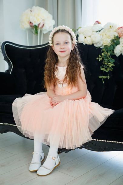 ein schönes kleines Mädchen in einem rosa Kleid und einem Blumenstrauß sitzt auf einem schwarzen Sofa. - Foto, Bild