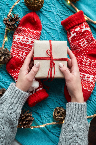 Mani femminili che tengono la scatola regalo di Natale sullo sfondo di calzini rossi, un maglione blu lavorato a maglia e una ghirlanda con coni
 - Foto, immagini