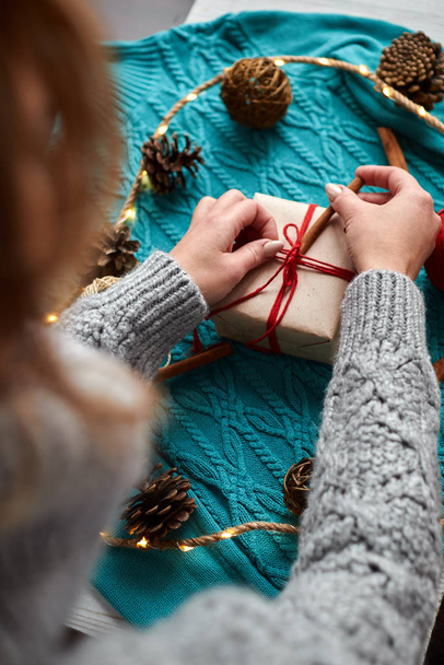 Руки женщины в пуловере держат рождественскую подарочную коробку, украшенную вечнозеленой веткой. Подготовка к декабрьским праздникам
 - Фото, изображение