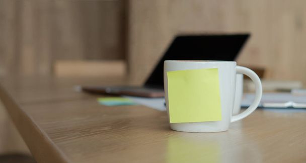 Valkoinen kahvikuppi, jossa on tyhjä keltainen Sticky Note. Lepää pöydällä ja kannettava tietokone on sijoitettu taakse
. - Valokuva, kuva