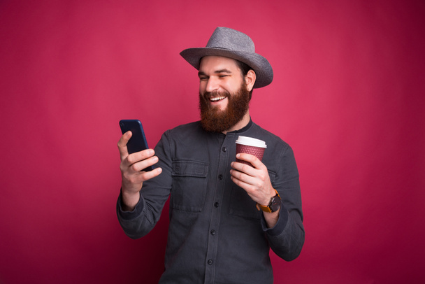 Foto de un joven sonriente con barba mirando el teléfono inteligente y sosteniendo una taza de café
 - Foto, Imagen