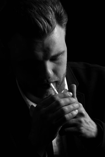 hipster young man smokes a cigarette. the guy is focused and serious on a dark background. black and white photo - Photo, image
