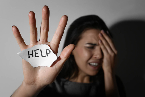 Crying young woman with sign HELP near white wall, focus on hand. Domestic violence concept - Foto, Imagen