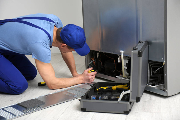 Male technician with screwdriver repairing refrigerator indoors - Foto, afbeelding