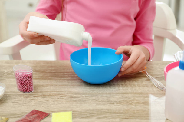 Little girl pouring glue into bowl at table in room, closeup. DIY slime toy - Φωτογραφία, εικόνα