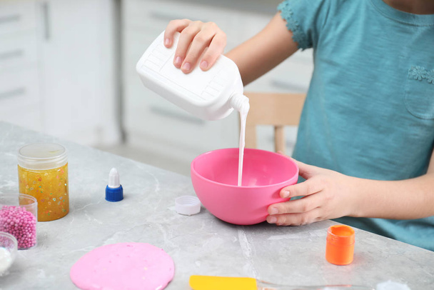 Little girl pouring glue into bowl at table in kitchen, closeup. DIY slime toy - Foto, Imagem