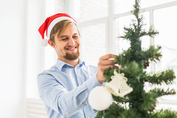 glücklicher Mann, der zu Hause den Weihnachtsbaum mit Weihnachtsmann-Hut schmückt. Mann schmückt Baum im Winterurlaub mit Christbaumkugeln. - Foto, Bild