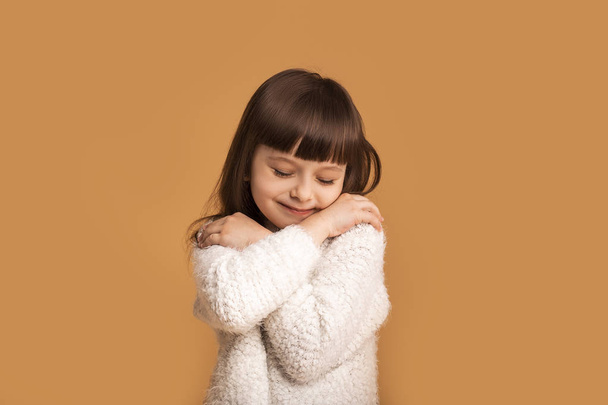 Αγάπη για τον εαυτό της, όμορφο κοριτσάκι που αγκαλιάζεται. Ζώνη άνεσης, απόλαυση, ζεστή έννοια. Χαρούμενο γοητευτικό καυκάσιο παιδί σε λευκό πουλόβερ πάνω από πορτοκαλί φόντο. - Φωτογραφία, εικόνα