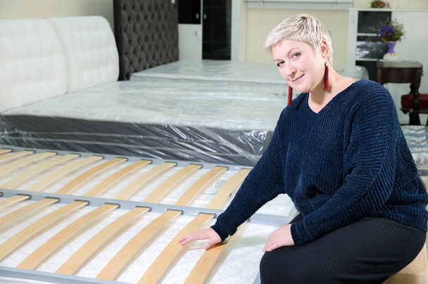 Stylish adult woman with short hair chooses an orthopedic bed in a furniture store. The concept of choice of health and orthopedic basis of a double bed - Foto, Bild