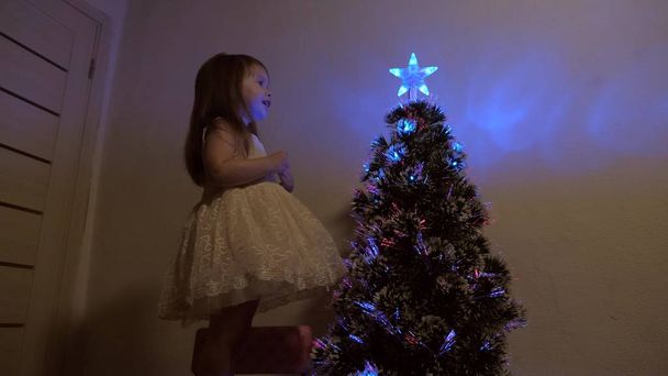 joyeux Noël concept de vacances pour les enfants. enfant examine une étoile de Noël sur un arbre de vacances. petite fille joue près d'un arbre de Noël dans une chambre d'enfant. magnifique arbre de Noël artificiel
. - Photo, image
