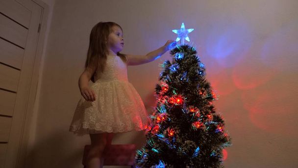 conceito de férias de Natal feliz para crianças. a criança examina uma estrela de Natal em uma árvore de férias. menina brinca perto de uma árvore de Natal em uma sala de crianças. bela árvore de Natal artificial
. - Foto, Imagem