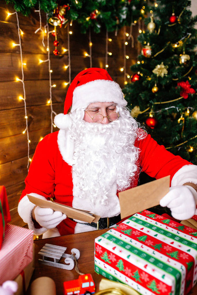 Merry Christmas and Happy Holidays. Santa Clause is preparing gifts for children for Xmas at desk at home. - Photo, image