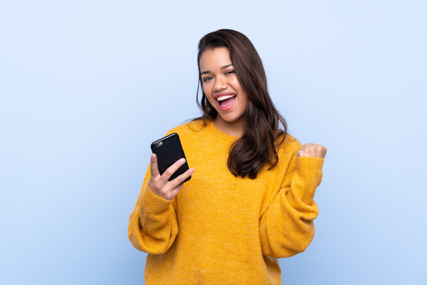 Mladá kolumbijská dívka se svetrem přes izolované modré pozadí s telefonem ve vítězné pozici - Fotografie, Obrázek