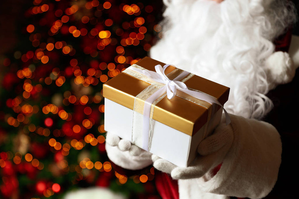 Santa Claus holding Christmas gift against blurred festive lights, closeup - Фото, изображение