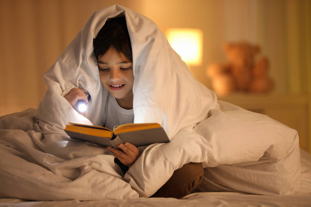 Chłopiec z latarką czyta książkę pod kocem w domu - Zdjęcie, obraz