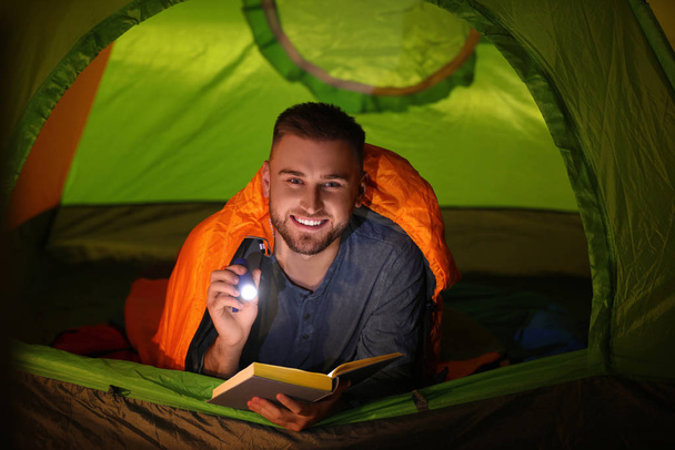 Young man with flashlight reading book in tent - Фото, изображение