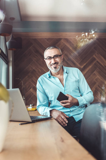 Muž středního věku v modré košili drží telefon vedle okna kanceláře. - Fotografie, Obrázek