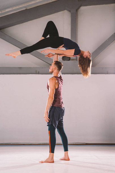 The theme of Acroyoga and Yoga Poses. A pair of two men and a woman stand in the position of asana. The guy holds the girl arched high back on the outstretched arm. In the gym with a studio Backlight - Фото, зображення