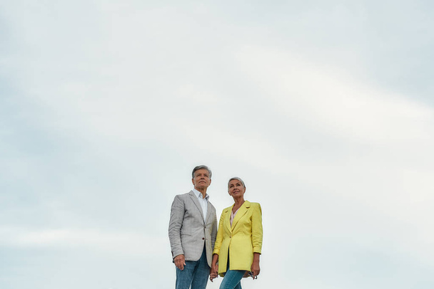 Just happy together. Elegant and lovely middle-aged couple holding hands and looking away while standing against blue sky - Fotografie, Obrázek