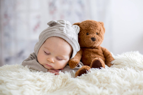 Sweet baby boy in bear overall, sleeping in bed with teddy bear - Фото, изображение