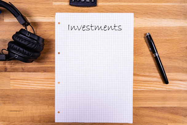 Блокнот с запиской "Инвестиции" вместе с ручкой, калькулятором и наушниками на деревянном столе
 - Фото, изображение