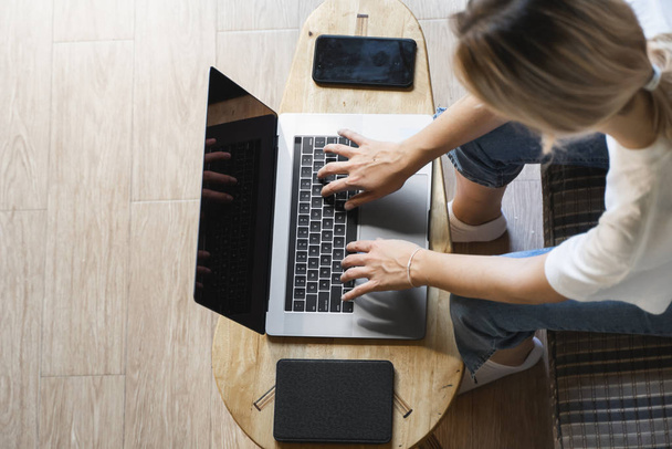 Vrouw zit op een bank en gebruikt een laptop aan houten tafel. Studeer en werk online, freelance. Zelfstandig meisje werkt met haar notebook zitten op een bank met een telefoon en ereader op tafel. - Foto, afbeelding