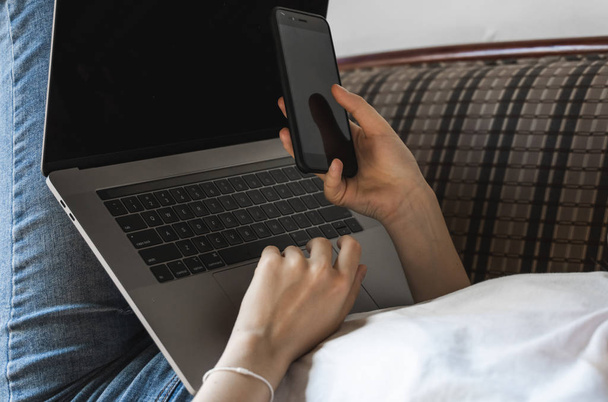 Woman with a laptop is laying on a sofa and using a smartphone. Study and work online, freelance. Self employed girl working with her notebook laying on a couch with a phone. - Photo, image
