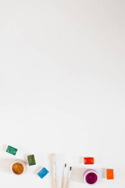 Кюветы с разноцветными акварельными красками и кисточками, банки с гуашами на белом фоне. Граница с инструментами рисования. Копирование текста
 - Фото, изображение