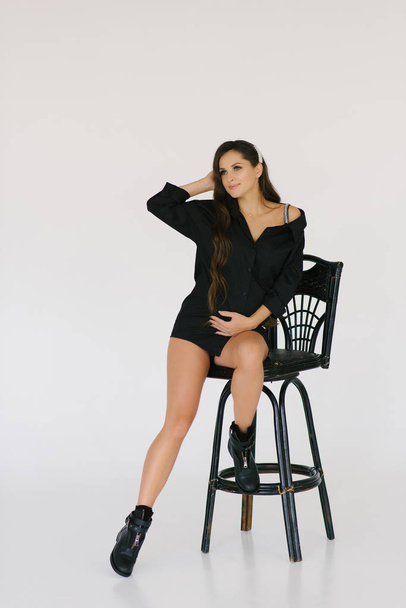 A beautiful long-haired brunette in a black shirt sits on a chair and holds her hand on her stomach. Happy waiting for baby's birth and healthy stylish pregnancy - Foto, Imagen