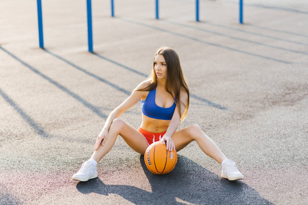 Hoikka kaunis tyttö urheiluunivormussa istuu leikkikentällä ja pitää koripalloa jalkojensa välissä. Koripalloilija
 - Valokuva, kuva
