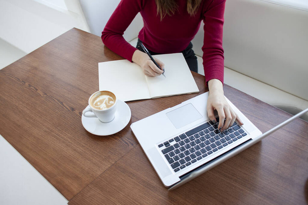 молодая девушка фрилансер сидит в кафе с ноутбуком и пишет в блокноте, студент учится за компьютером утром
 - Фото, изображение