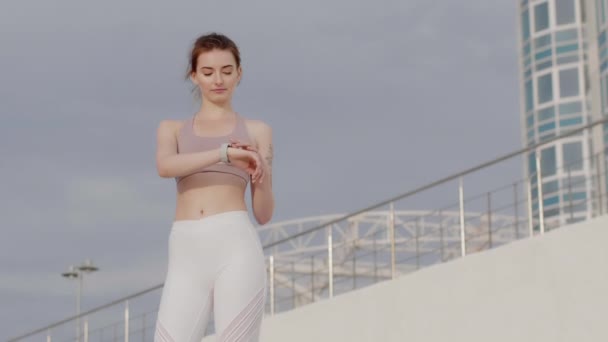 Jovem mulher atraente no topo desportivo confiantemente verificando relógio inteligente após o treino na praia
 - Filmagem, Vídeo