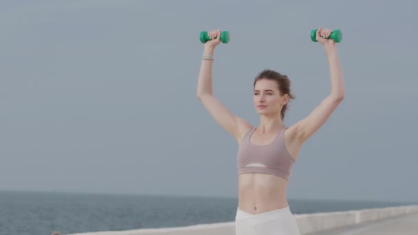Joven hermosa mujer deportiva haciendo ejercicios de mano con pesas durante el entrenamiento junto al mar
 - Metraje, vídeo