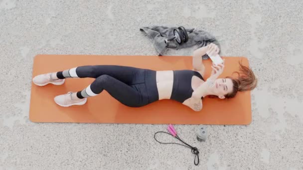 Bovenaanzicht van jonge vrij sportieve vrouw liggend op yoga mat gelukkig het nemen van selfie op mobiele telefoon na training outdoor - Video