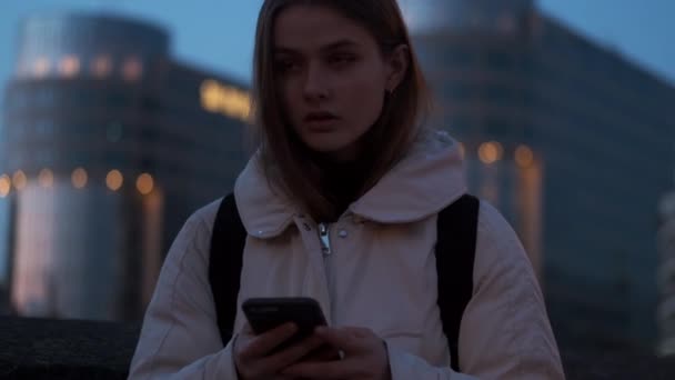 Μέτριο πλάνο του ελκυστικού casual κορίτσι σκεπτικά κοιτάζοντας κατά μέρος χρησιμοποιώντας το κινητό τηλέφωνο στο δρόμο βράδυ το σούρουπο - Πλάνα, βίντεο