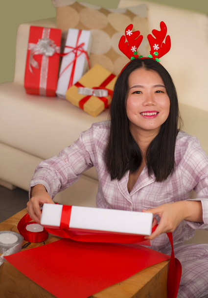 Christmas holidays lifestyle portrait of young beautiful and happy Asian Chinese woman in reindeer horns hat excited wrapping presents packing gifts with paper - Photo, Image