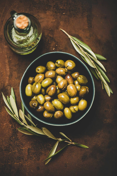 Flat-lay of fresh harvested seasonal pickled green Mediterranean olives in bowl and olive oil in bottle over dark rusty background, top view. Healthy snack, starter or traditional meze food - Fotografie, Obrázek