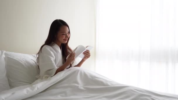 footage of beautiful asian woman in bathrobe reading book in bed - Filmagem, Vídeo