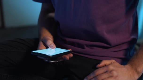 Зрілий хлопець у фіолетовій футболці сидить сам у темній кімнаті і використовує свій смартфон
. - Кадри, відео