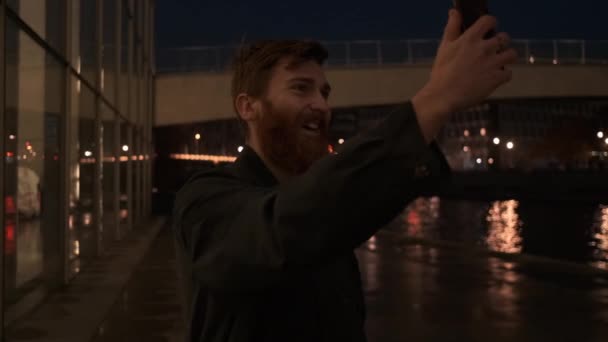 Sledování kolem záběru mladého atraktivní vousatý muž šťastně mluví na video chat na mobilu ve městě ulici v noci - Záběry, video