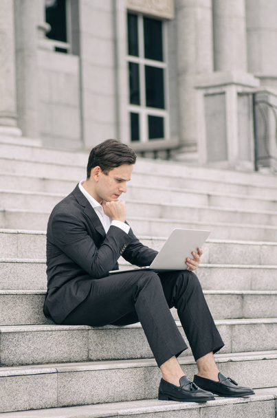 разочарованный и подавленный молодой бизнесмен сидит на лестнице, работая со своим ноутбуком
 - Фото, изображение