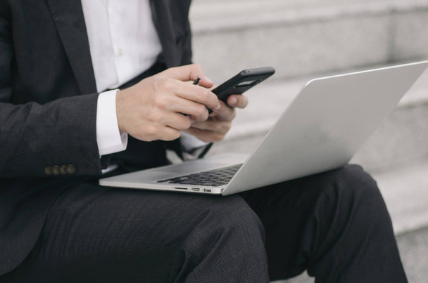 ongeïdentificeerde jonge zakenman in elegant pak met laptop, zittend en met behulp van zijn mobiele telefoon - Foto, afbeelding