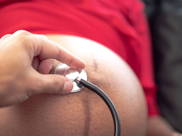Een close-up van de hand van een mannelijke arts of echtgenoot die een stethoscoop op de blote buik of buik van een zwangere vrouw of vrouw met een rode jurk draagt om te luisteren naar de hartslag van de ongeboren baby. - Foto, afbeelding